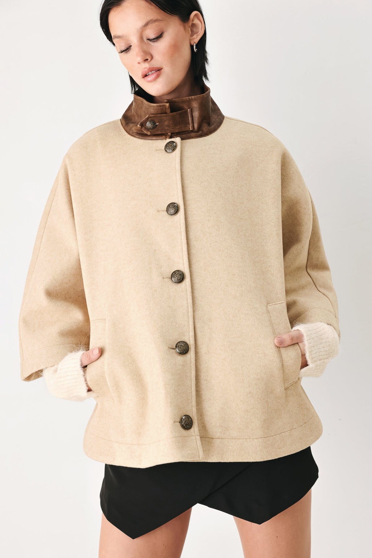 A woman wearing a beige wool-blend coat with a contrasting brown collar, dark brown buttons, and ribbed cream cuffs, paired with black shorts, against a plain light gray background.