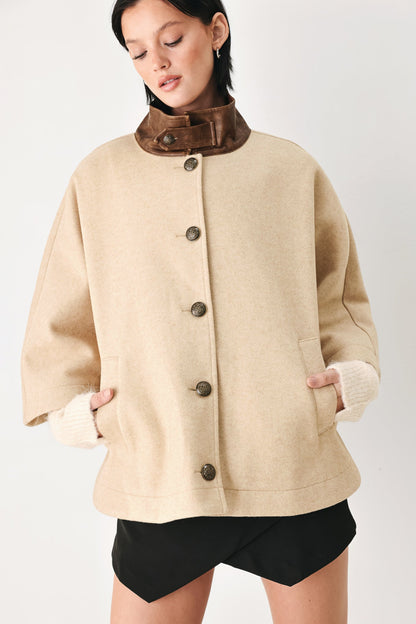 A woman wearing a beige wool-blend coat with a contrasting brown collar, dark brown buttons, and ribbed cream cuffs, paired with black shorts, against a plain light gray background.