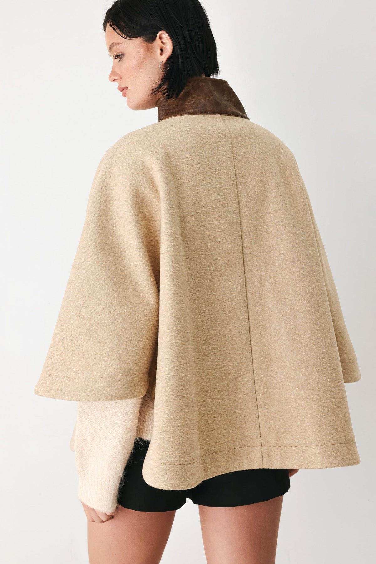 A woman shown from the back wearing a beige cape-style coat with a dark brown collar, paired with black shorts, against a plain light background.