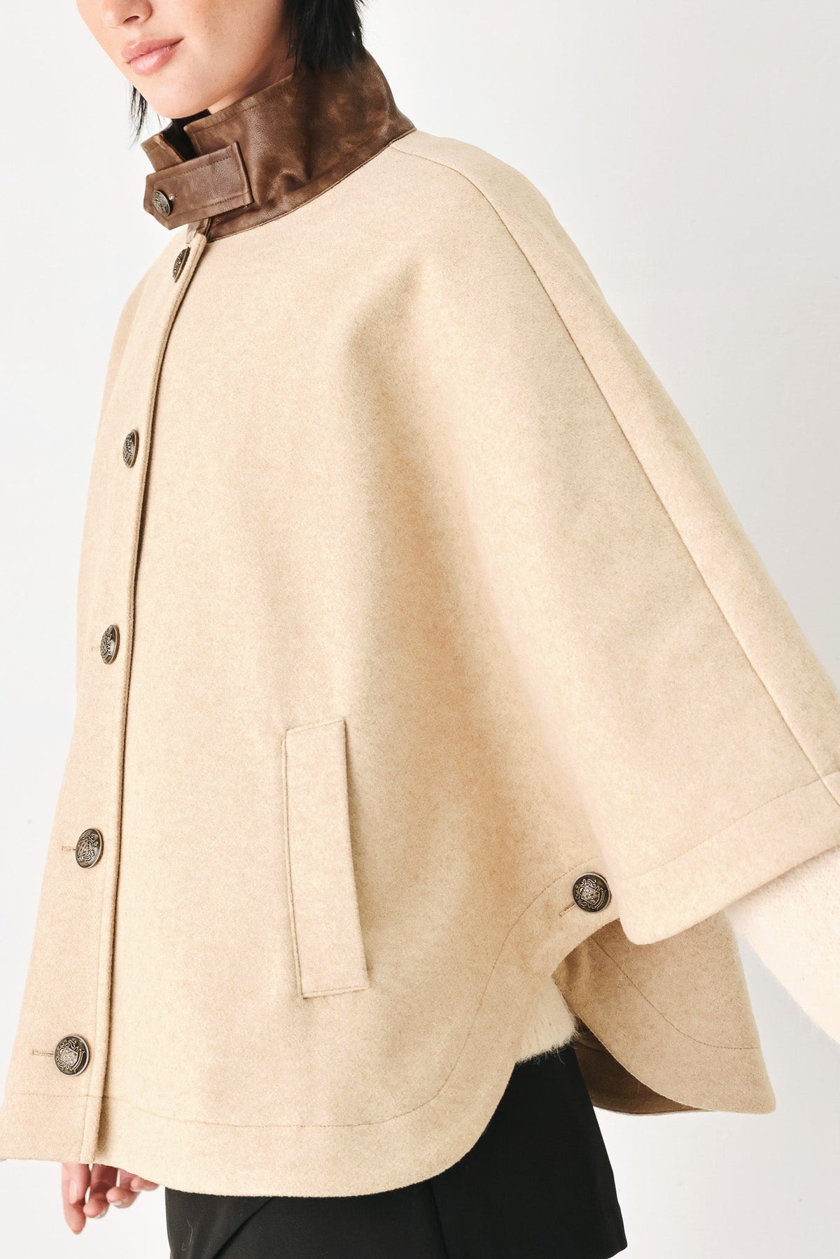 A close-up of a model wearing a beige cape-style coat with a brown collar and dark buttons, set against a plain light background.