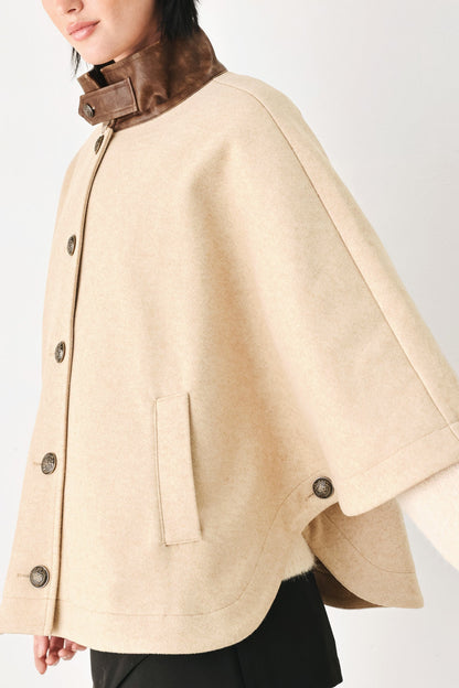 A close-up of a model wearing a beige cape-style coat with a brown collar and dark buttons, set against a plain light background.