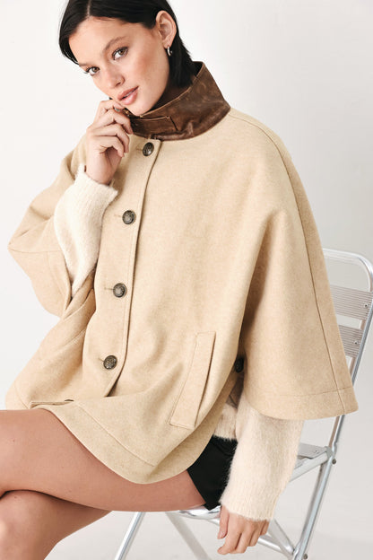A woman seated on a metal chair wearing a beige cape-style coat with a brown collar and dark buttons, over a cream knit sweater.