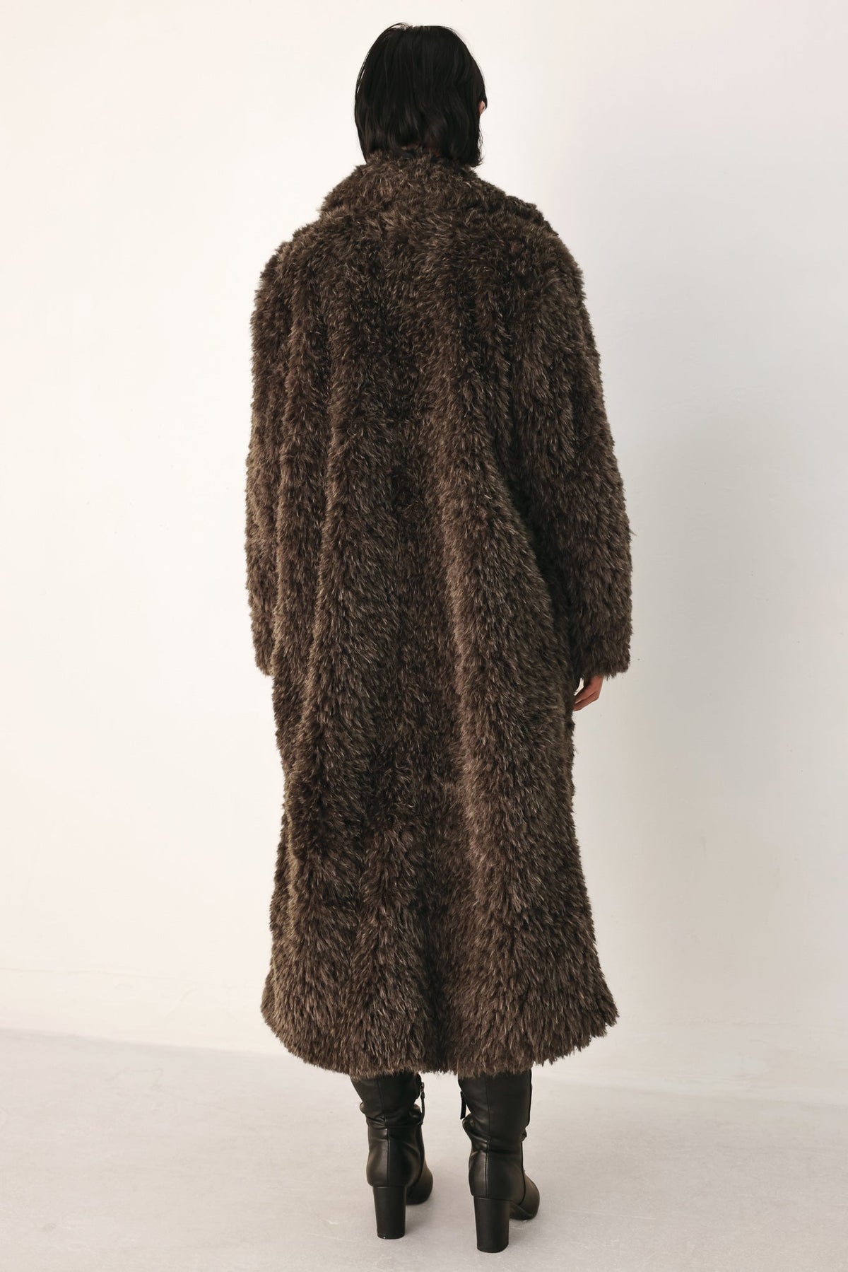 A person viewed from the back wearing a long, shaggy brown faux-fur coat over black knee-high boots, standing against a plain light background.