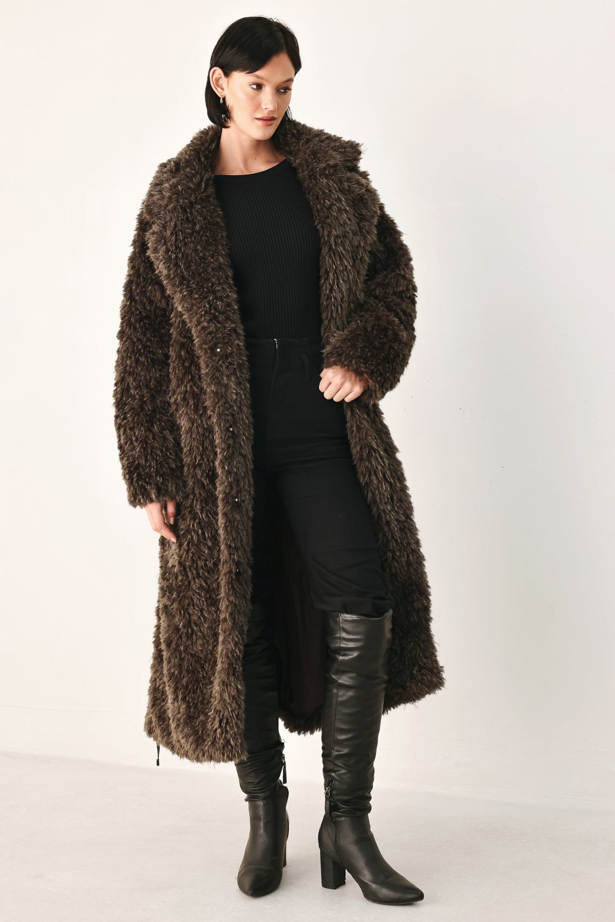 A woman modeling a long, dark brown shaggy faux-fur coat over a black outfit and black knee-high boots, photographed against a plain light studio background.