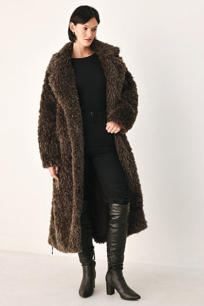 A woman modeling a long, dark brown shaggy faux-fur coat over a black outfit and black knee-high boots, photographed against a plain light studio background.
