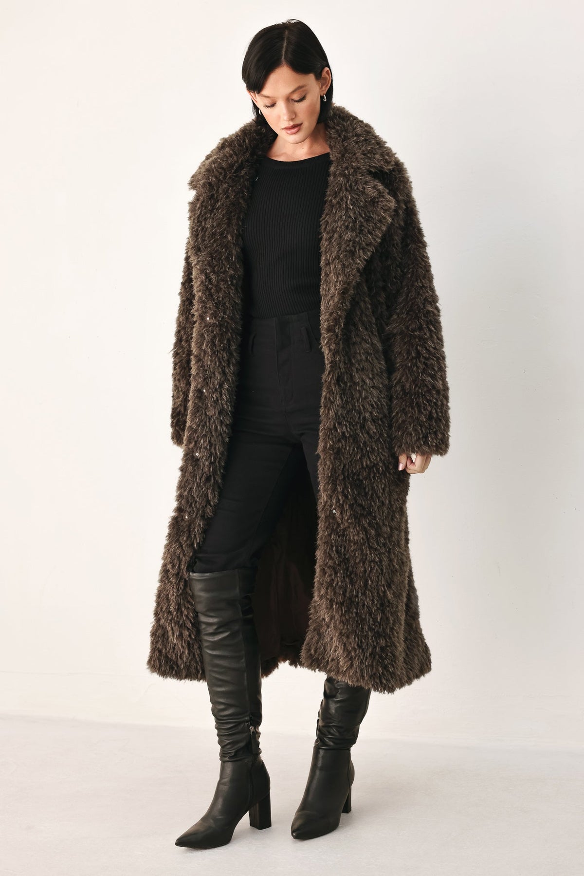 A model wearing a long, dark brown shaggy faux fur coat over a black outfit and black knee-high boots, standing against a plain off-white studio background.