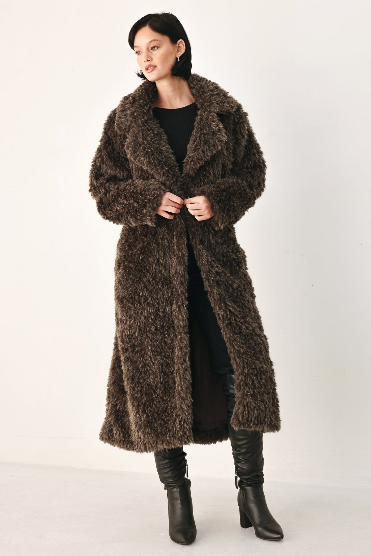 A woman modeling a long, shaggy brown faux fur coat over a black outfit and black knee-high boots, photographed against a plain light studio background.