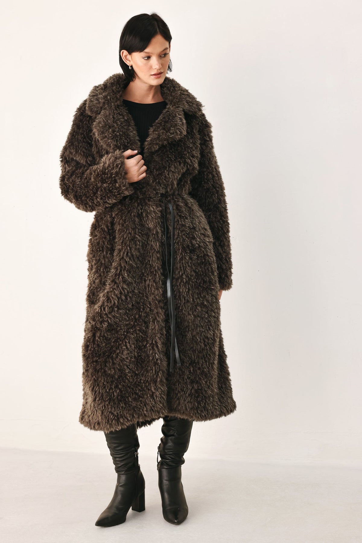 A model wearing a long, dark brown shaggy faux fur coat over a black top and black leather boots, standing against a plain off-white studio background.
