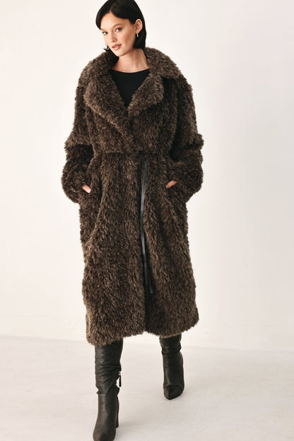 A woman modeling a long, shaggy brown faux fur coat with a wide collar, worn over a black top and black knee-high boots, against a plain white studio background.