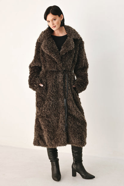 A woman modeling a long, shaggy brown faux fur coat with a black top and black heeled boots, standing against a plain light gray studio background.