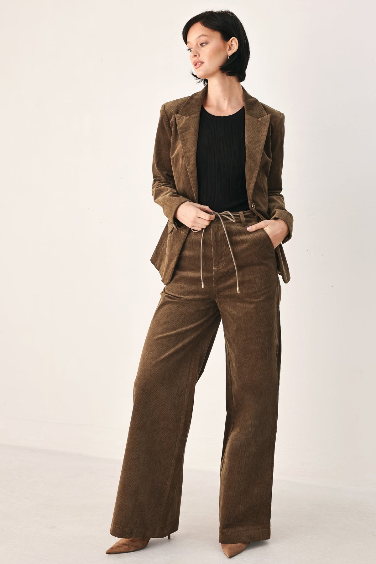 A model wearing a matching brown corduroy blazer and wide-leg trousers set with a drawstring waist, styled over a black top and brown pointed-toe heels, against a plain off-white studio background.