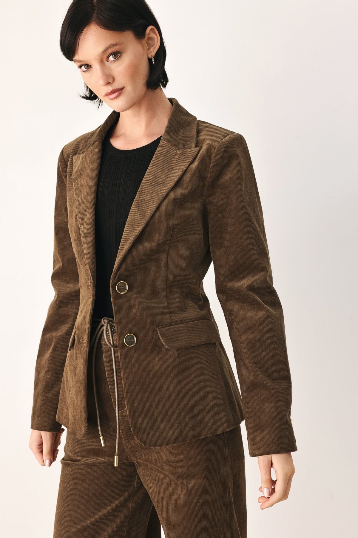 A woman modeling a brown suede-like blazer and matching pants set with a drawstring waist, worn over a black top against a plain light background.