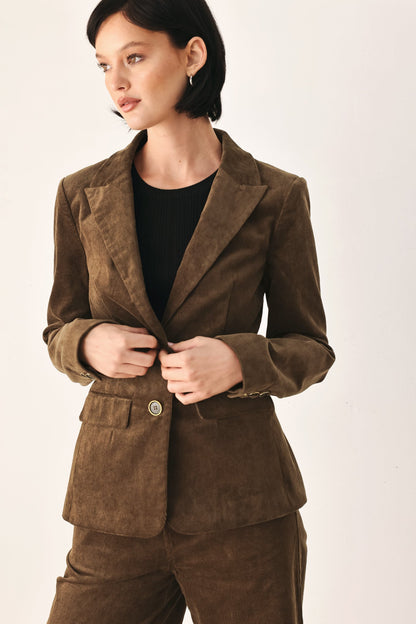 A woman wearing a brown corduroy blazer and matching trousers over a black top, photographed against a plain light background.