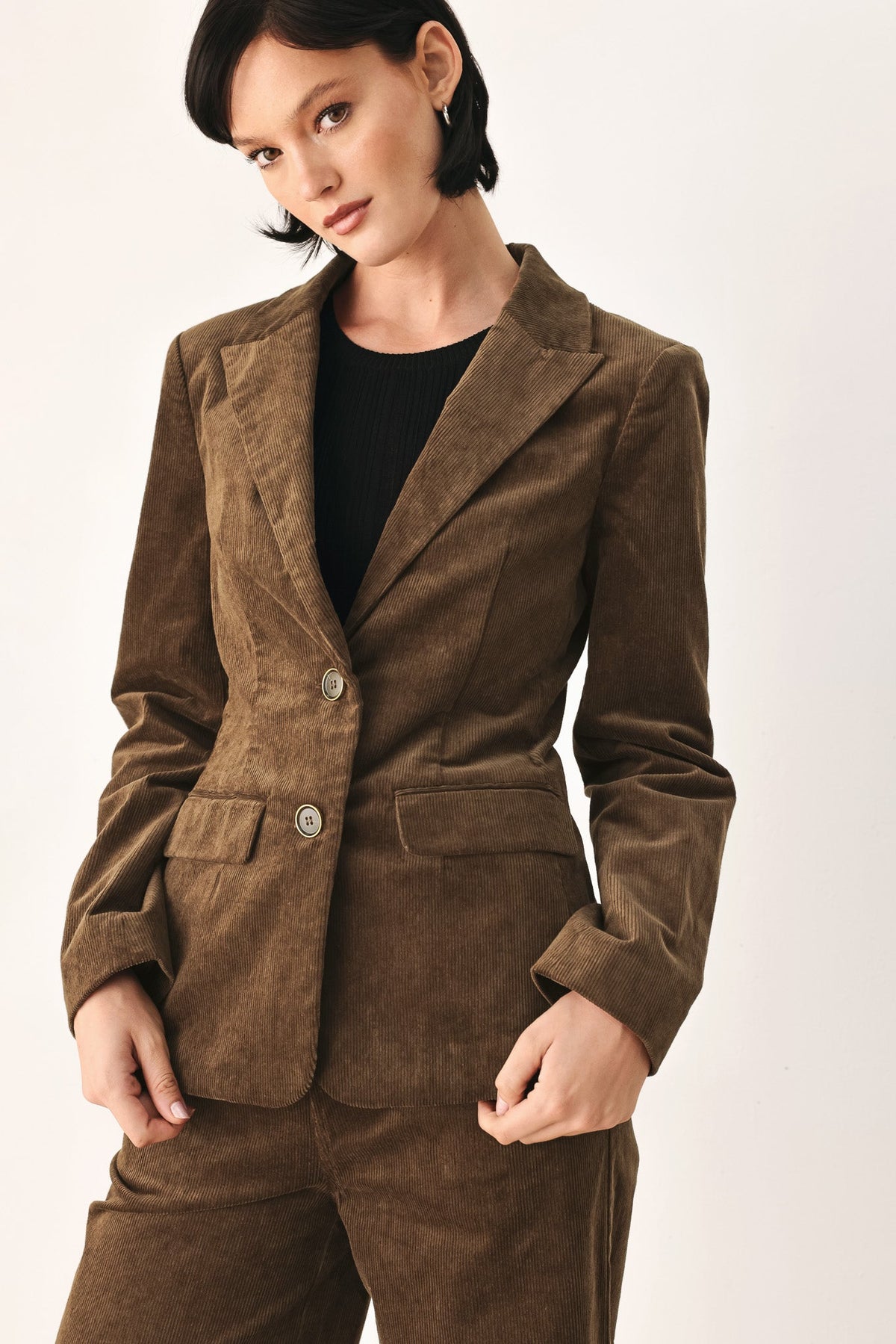 A woman wearing a brown corduroy blazer and matching trousers over a black top, photographed against a plain light background.