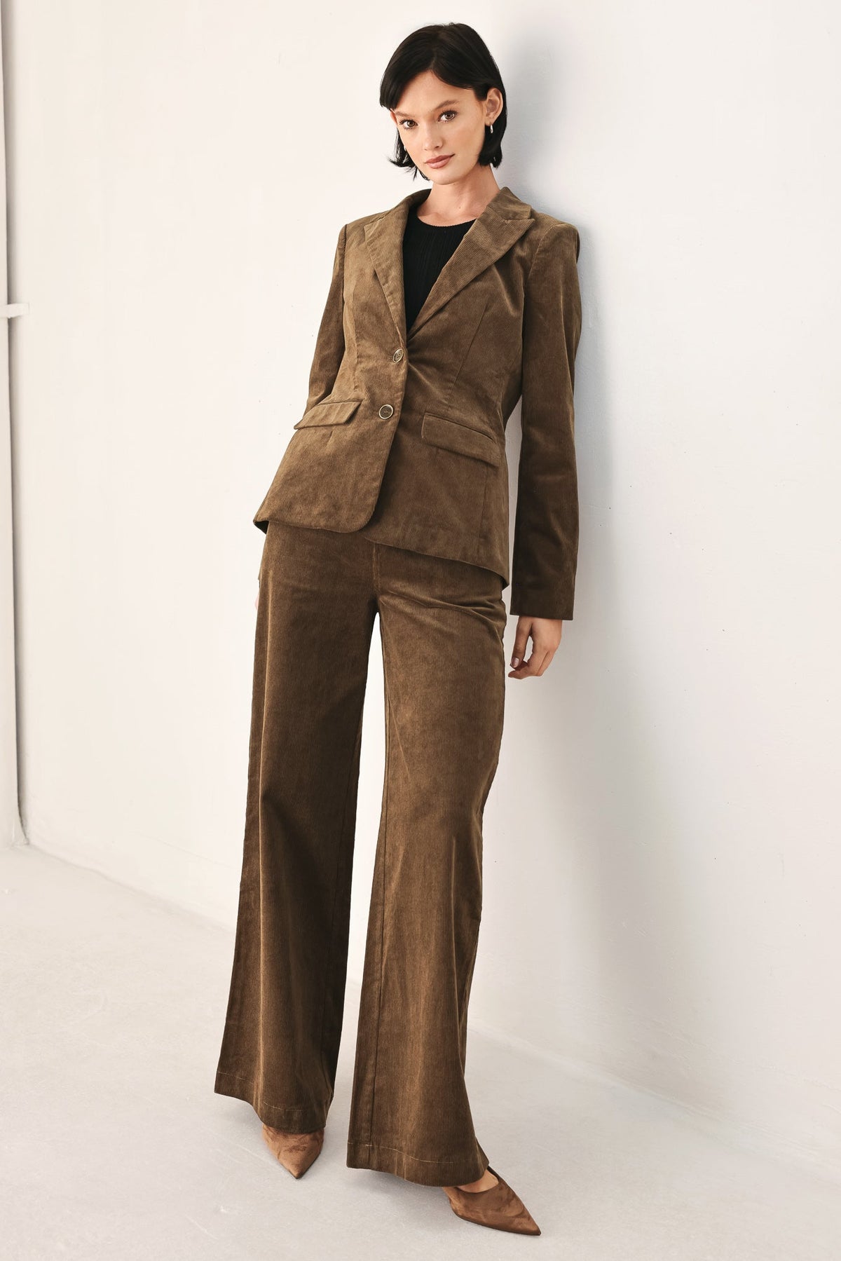 A model wearing a brown corduroy pantsuit consisting of a single-breasted blazer and matching wide-leg trousers, styled with a black top and brown pointed-toe flats against a white studio background.
