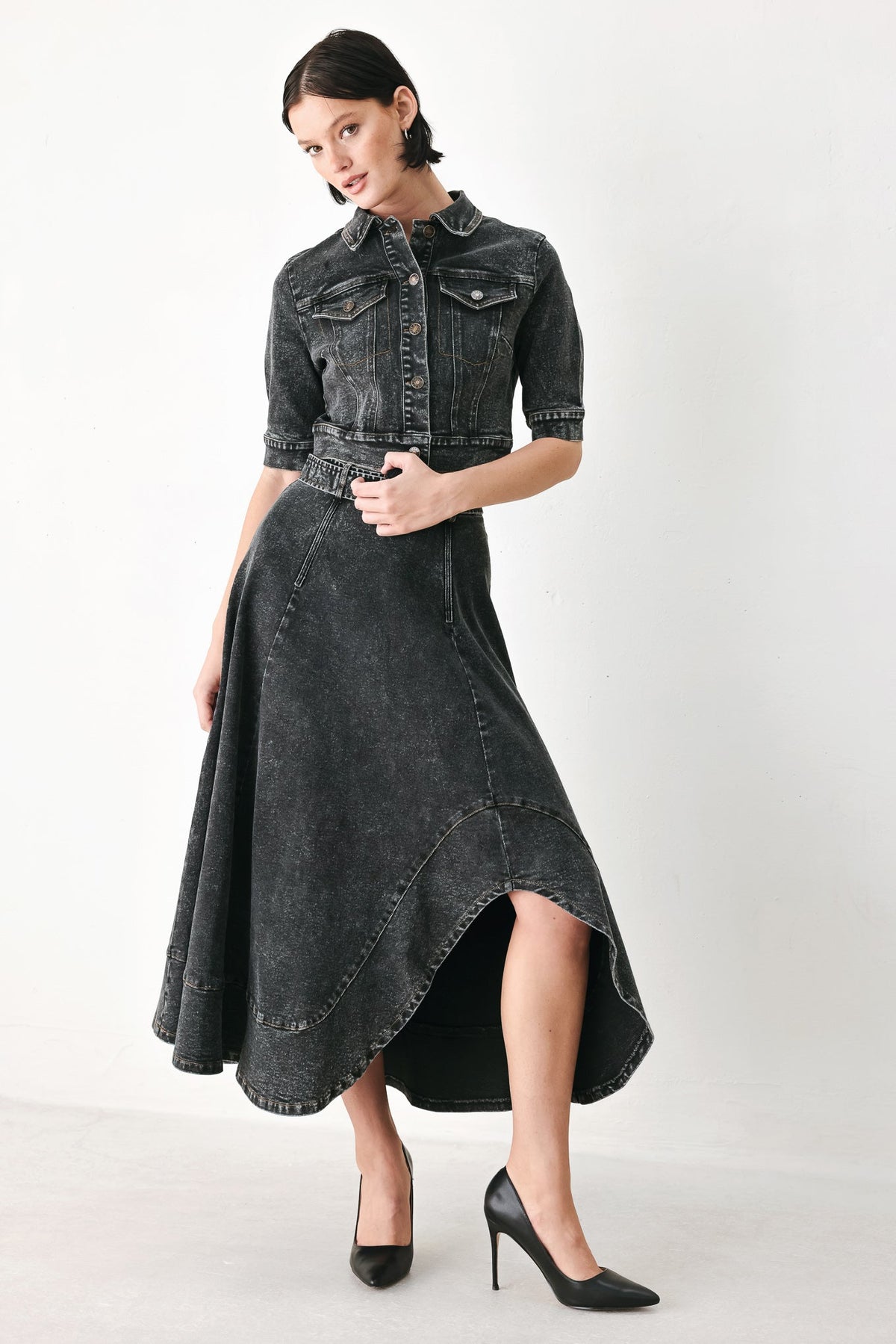 A model wearing a dark gray washed denim high-low dress with a button-up shirt collar, short sleeves, and a matching belt, paired with black heels, against a plain light gray background.