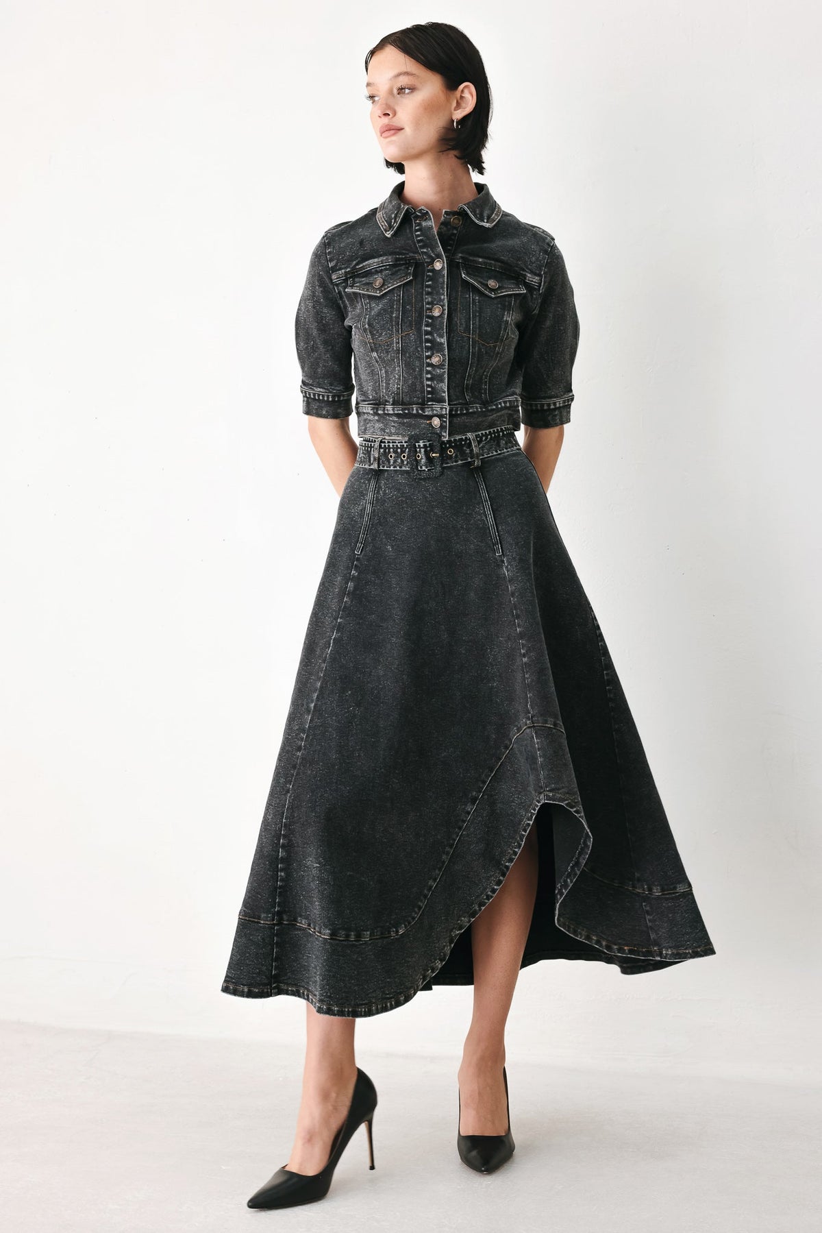 A model wearing a dark gray washed denim shirt-jacket and matching high-low skirt set, paired with black pointed-toe heels, against a plain white studio background.