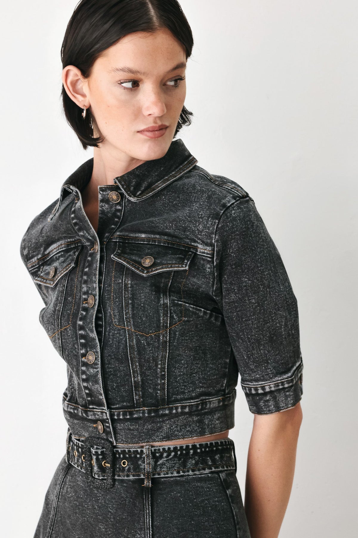 A model wearing a cropped, short-sleeved black acid-wash denim jacket with a band collar and button front, paired with matching studded denim bottoms, against a plain light gray background.