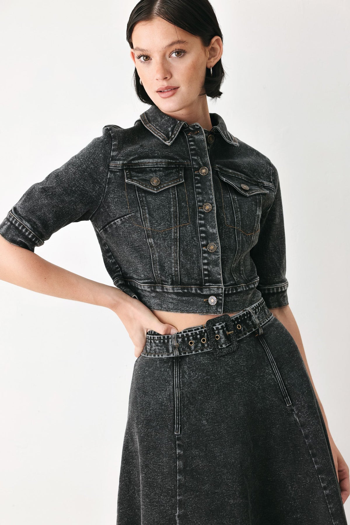 A model wearing a dark gray acid-wash denim jacket with rolled sleeves and a matching high-waisted denim skirt, shown against a plain white studio background.