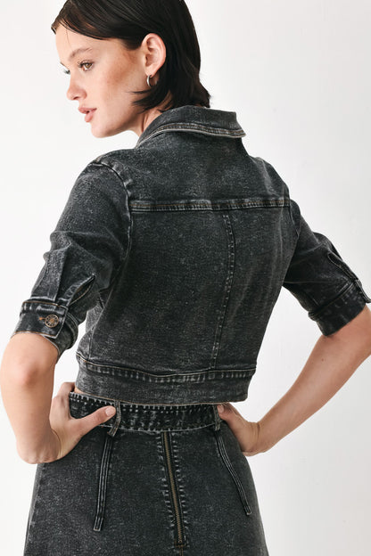 A model shown from the back wearing a cropped, acid-washed black denim jacket with rolled sleeves and a matching dark denim skirt against a plain white studio background.