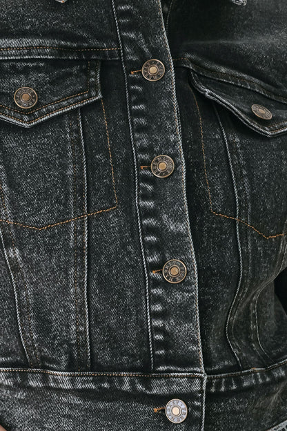 Close-up of a dark washed denim jacket showing the front button closure, chest pockets, and detailed denim texture.