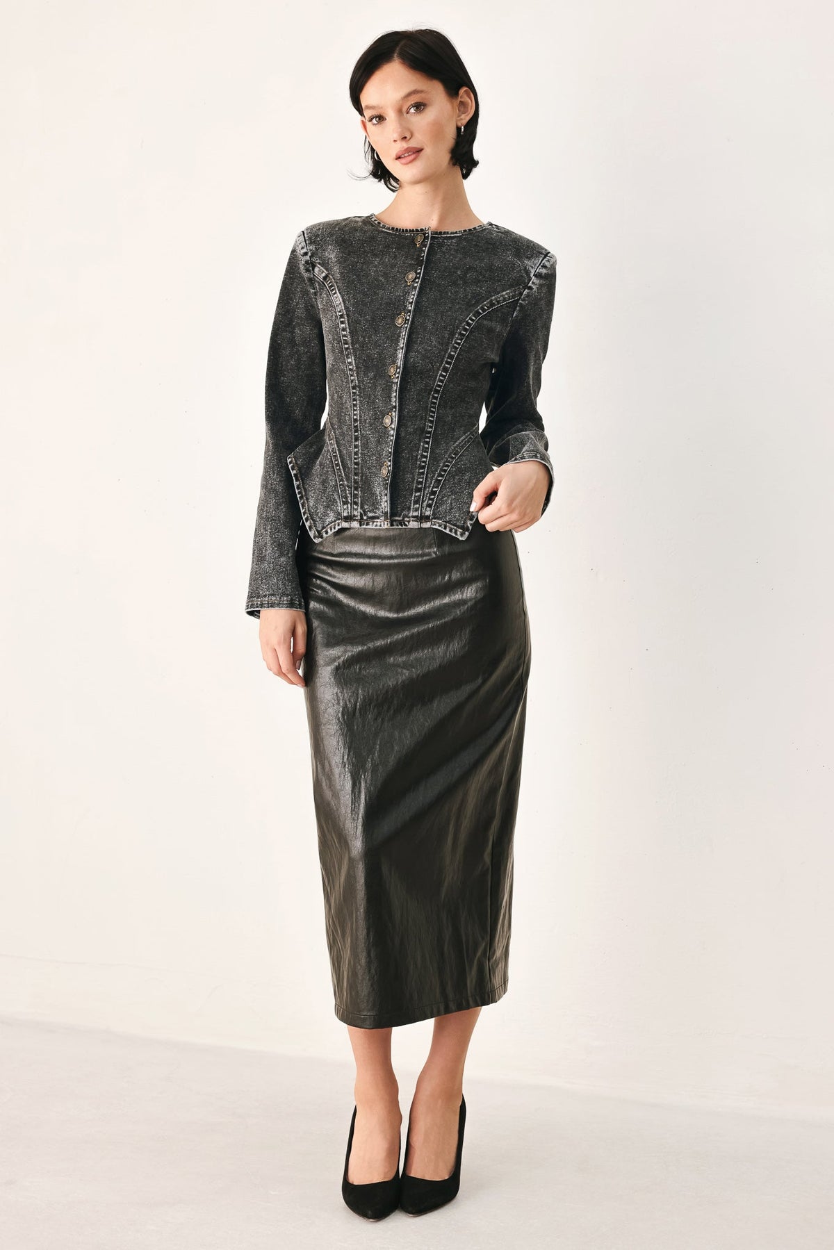 A model stands against a plain light background wearing a dark gray textured jacket with button details and a matching dark gray leather skirt, paired with black heels.