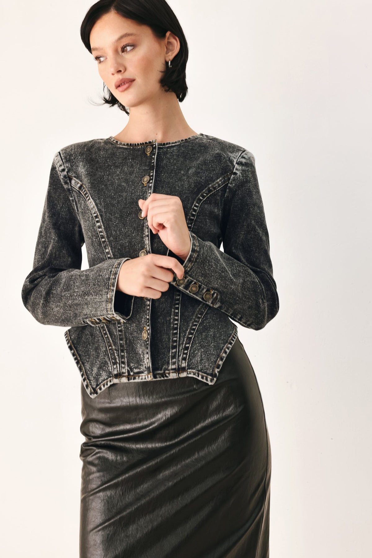 A woman wearing a cropped, long-sleeve dark gray acid-wash denim jacket with gold-tone buttons and a black faux-leather pencil skirt, photographed against a plain light background.