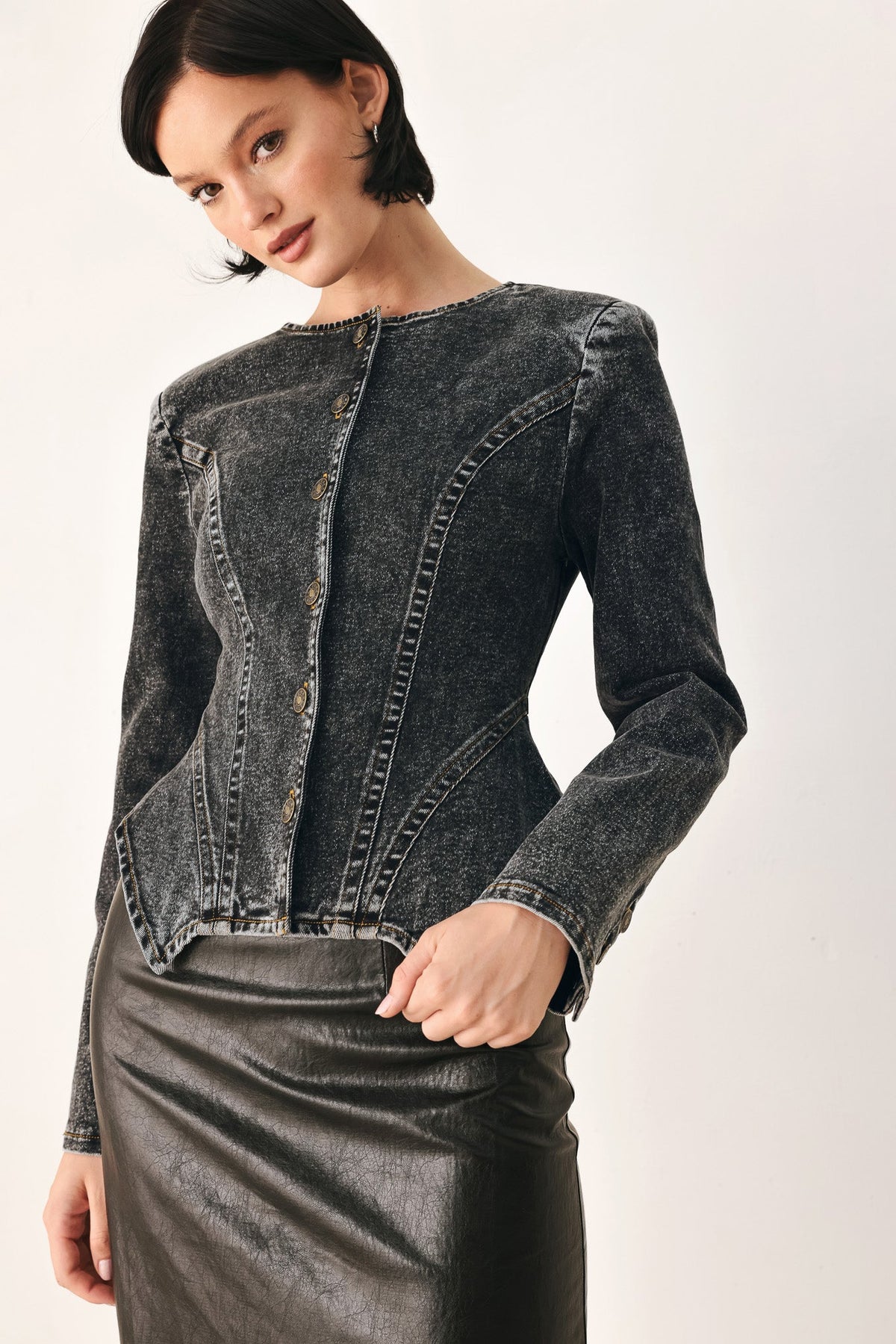 A model wearing a dark gray washed denim jacket with decorative button details down the front and a black leather skirt, photographed against a plain off-white studio background.