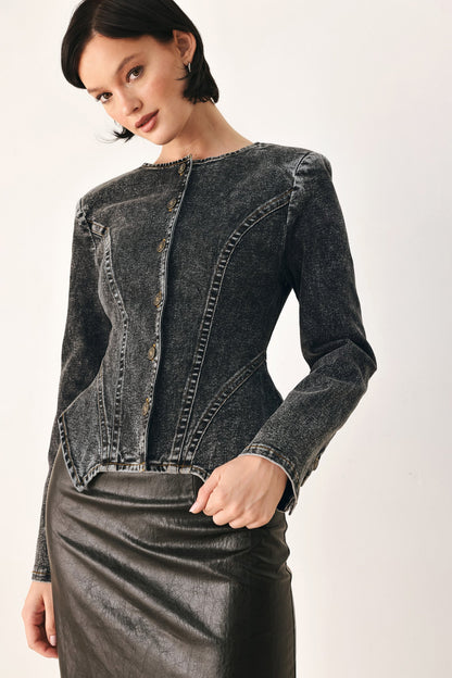 A model wearing a dark gray washed denim jacket with decorative button details down the front and a black leather skirt, photographed against a plain off-white studio background.