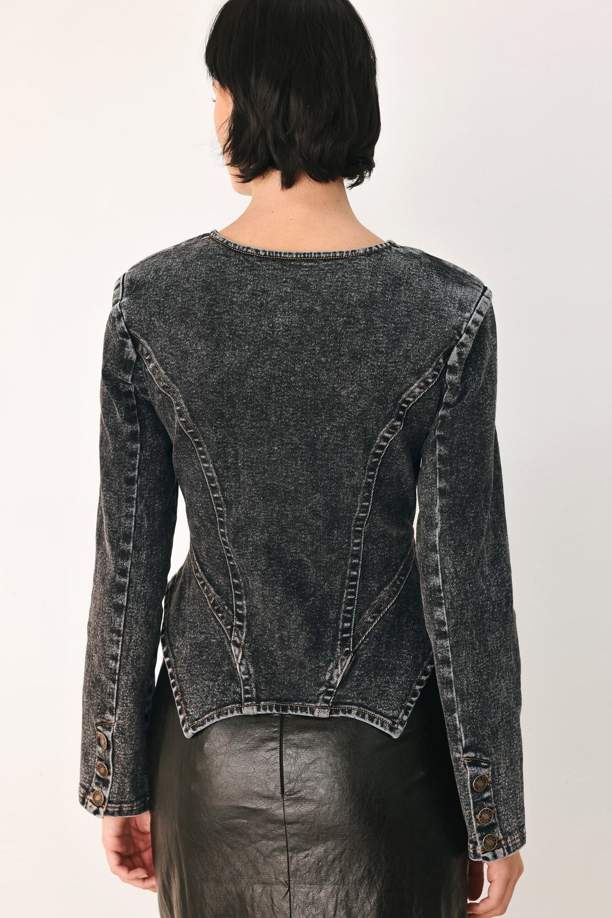 A model shown from the back wearing a dark gray acid-washed denim jacket with structured shoulders and a cropped fit, paired with a black leather skirt against a plain white studio background.
