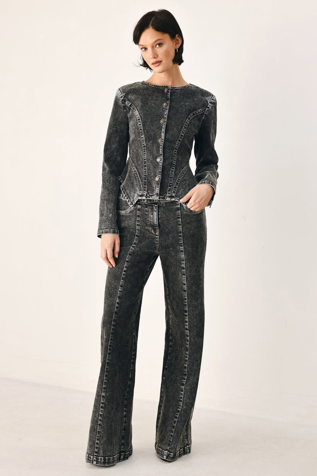 A model wearing a dark gray acid-washed denim jumpsuit with a button-up front and wide-leg pants, photographed against a clean off-white studio background.