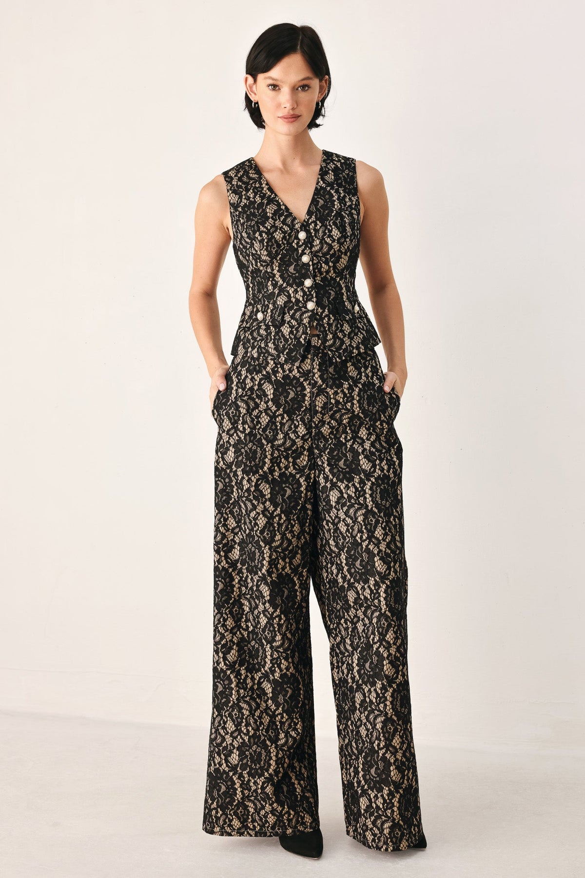A woman modeling a sleeveless black lace jumpsuit with a V-neckline, front buttons, and wide legs, photographed against a plain light studio background.