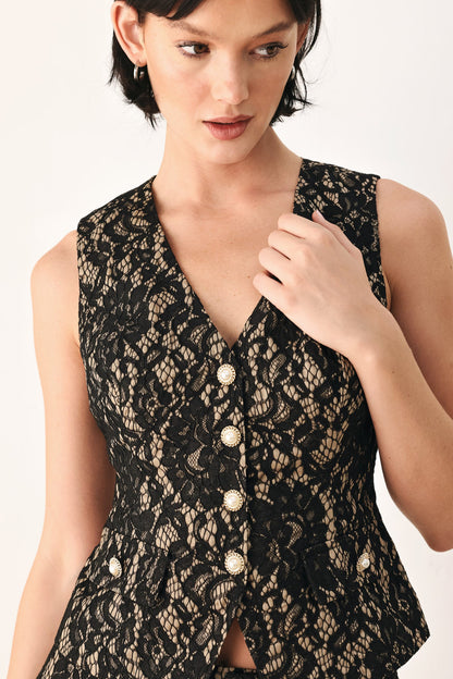 A close-up of a model wearing a sleeveless black lace vest with a V-neckline and decorative gold-tone buttons down the front, set against a plain light background.