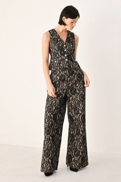 A woman modeling a sleeveless black lace jumpsuit with a buttoned V-neck bodice and wide legs, photographed against a plain light studio background.