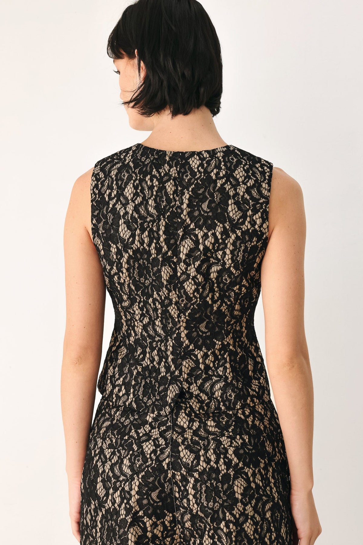 A woman shown from the back wearing a sleeveless black lace dress against a plain light background.