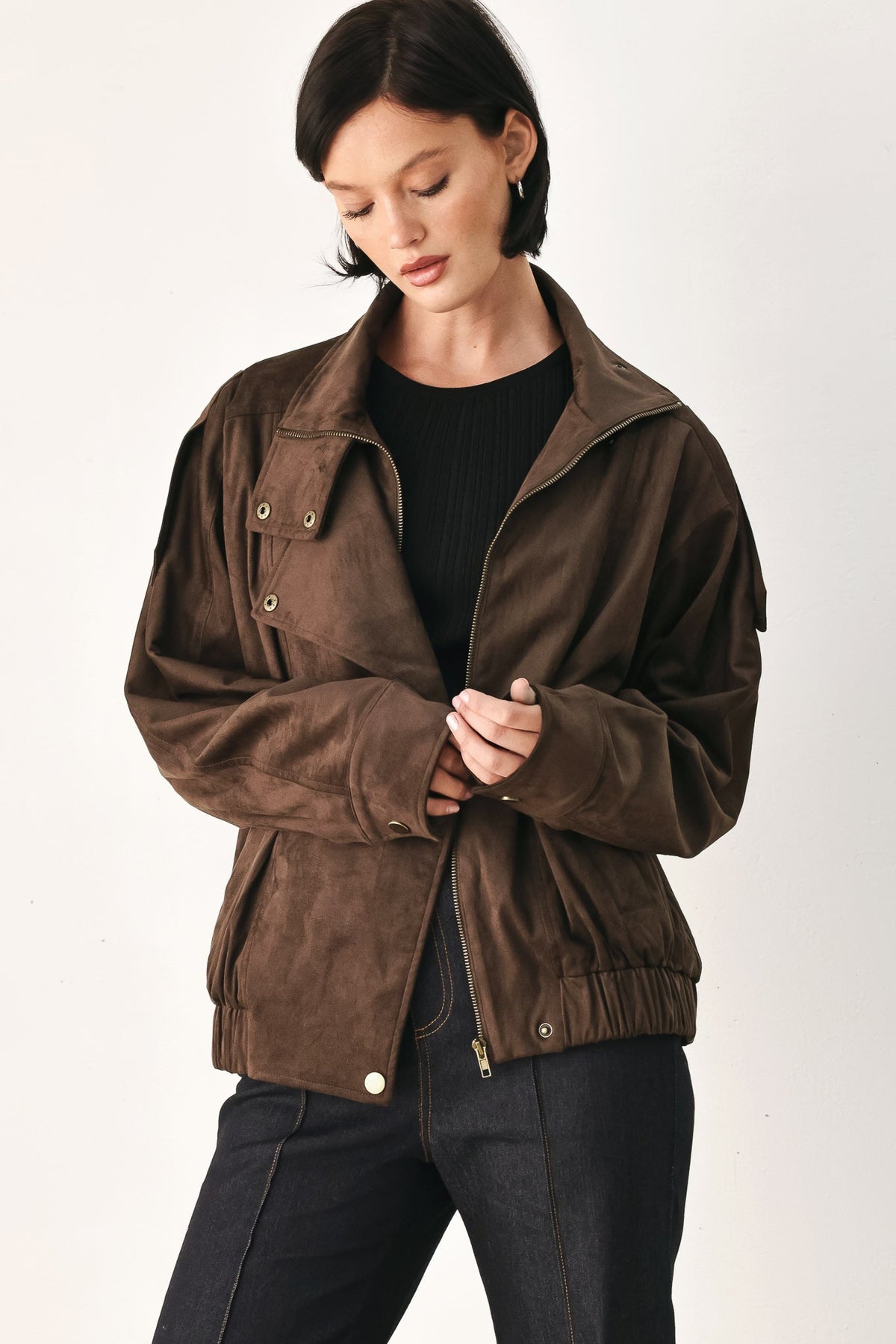 A woman wearing a brown suede utility jacket over a black top and dark jeans, photographed against a plain light background.