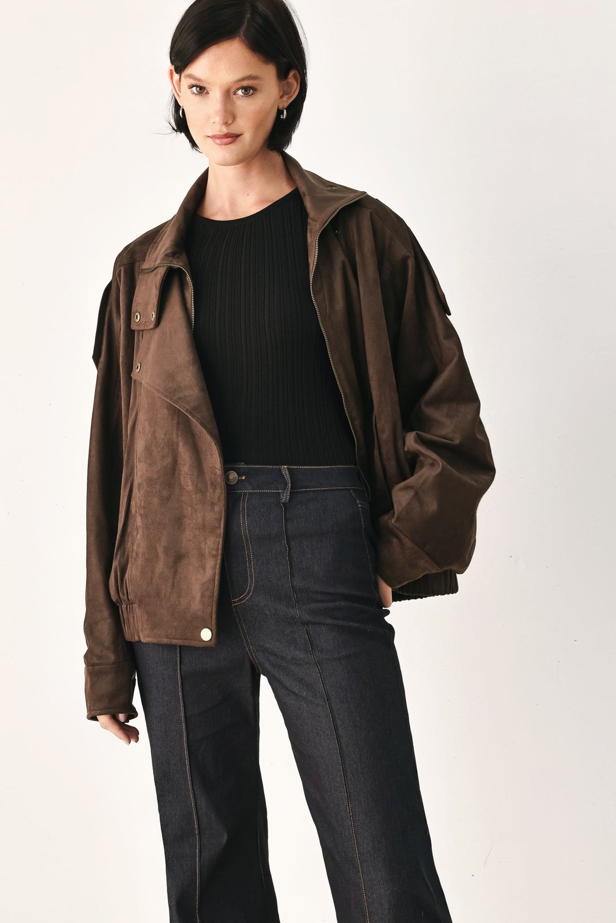 A model wearing a dark brown suede jacket over a black ribbed top and dark blue jeans, photographed against a plain off-white studio background.