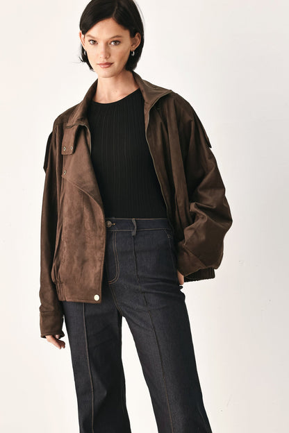 A model wearing a dark brown suede jacket over a black ribbed top and dark blue jeans, photographed against a plain off-white studio background.