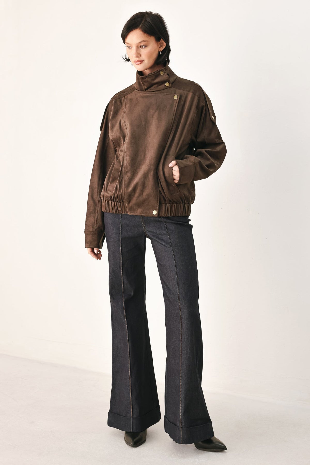 A model wearing a brown suede-like jacket with a high collar and dark wide-leg jeans, standing against a plain off-white studio background.