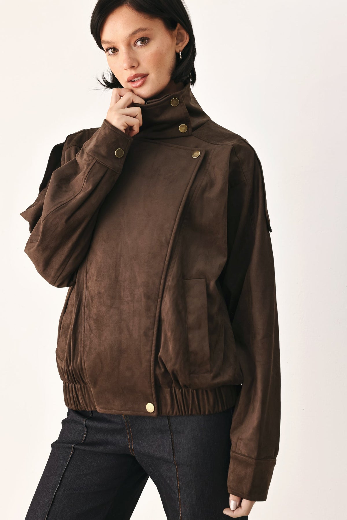 A woman wearing a dark brown suede jacket with a high collar and gold-tone snap buttons, paired with dark jeans, against a plain off-white studio background.