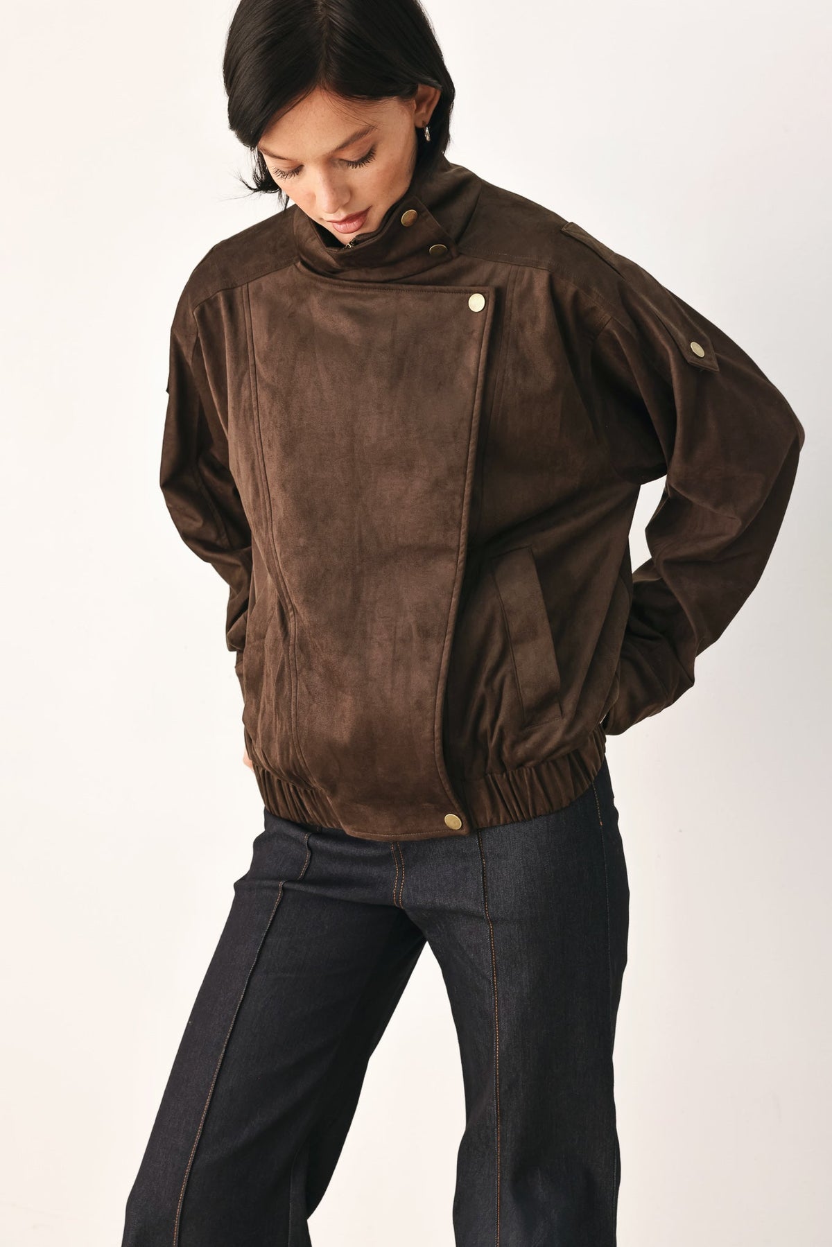 A model wearing a dark brown suede jacket with an asymmetrical snap closure and dark blue jeans, photographed against a plain light background.