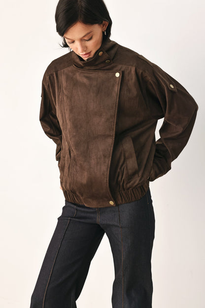 A model wearing a dark brown suede jacket with an asymmetrical snap closure and dark blue jeans, photographed against a plain light background.