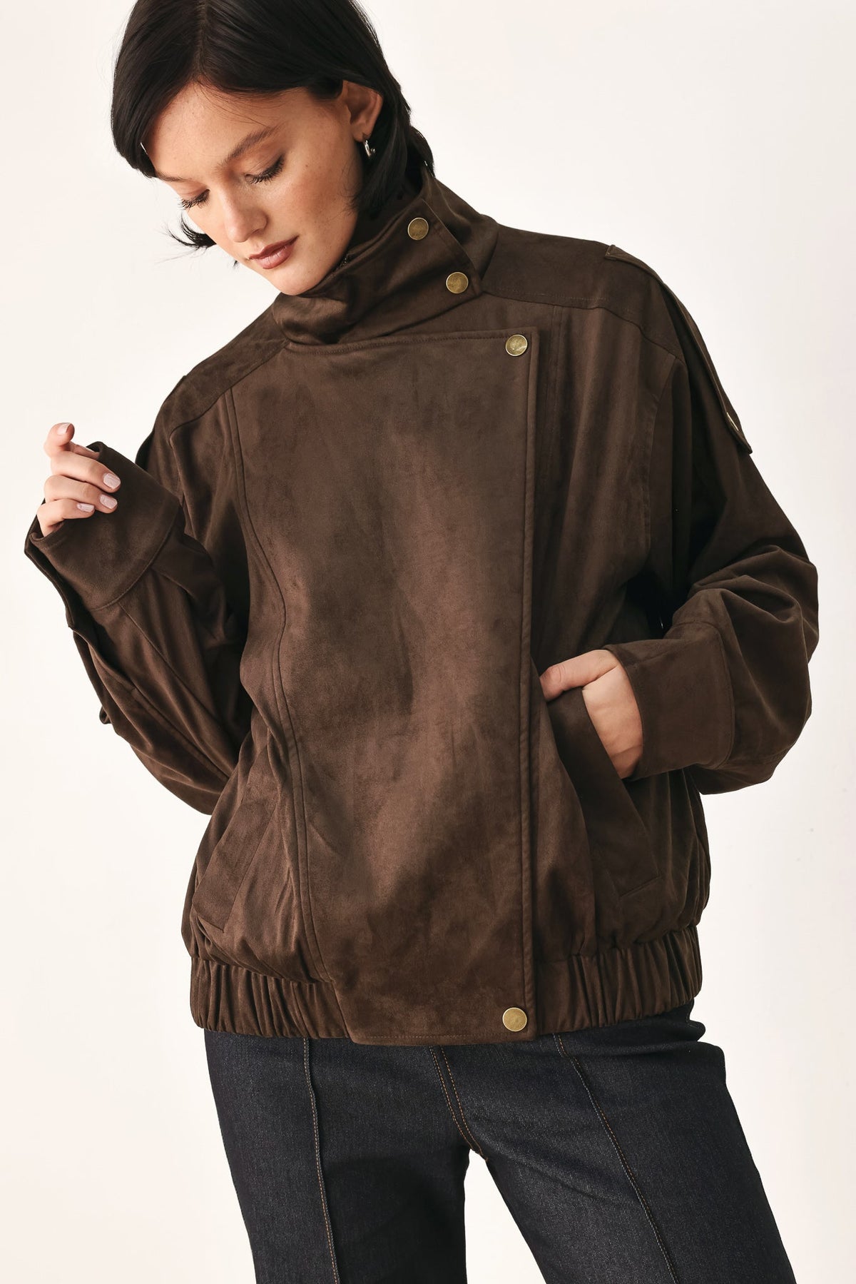 A woman wearing a dark brown suede jacket with gold-tone snap buttons and a high collar, paired with dark jeans, photographed against a plain light background.