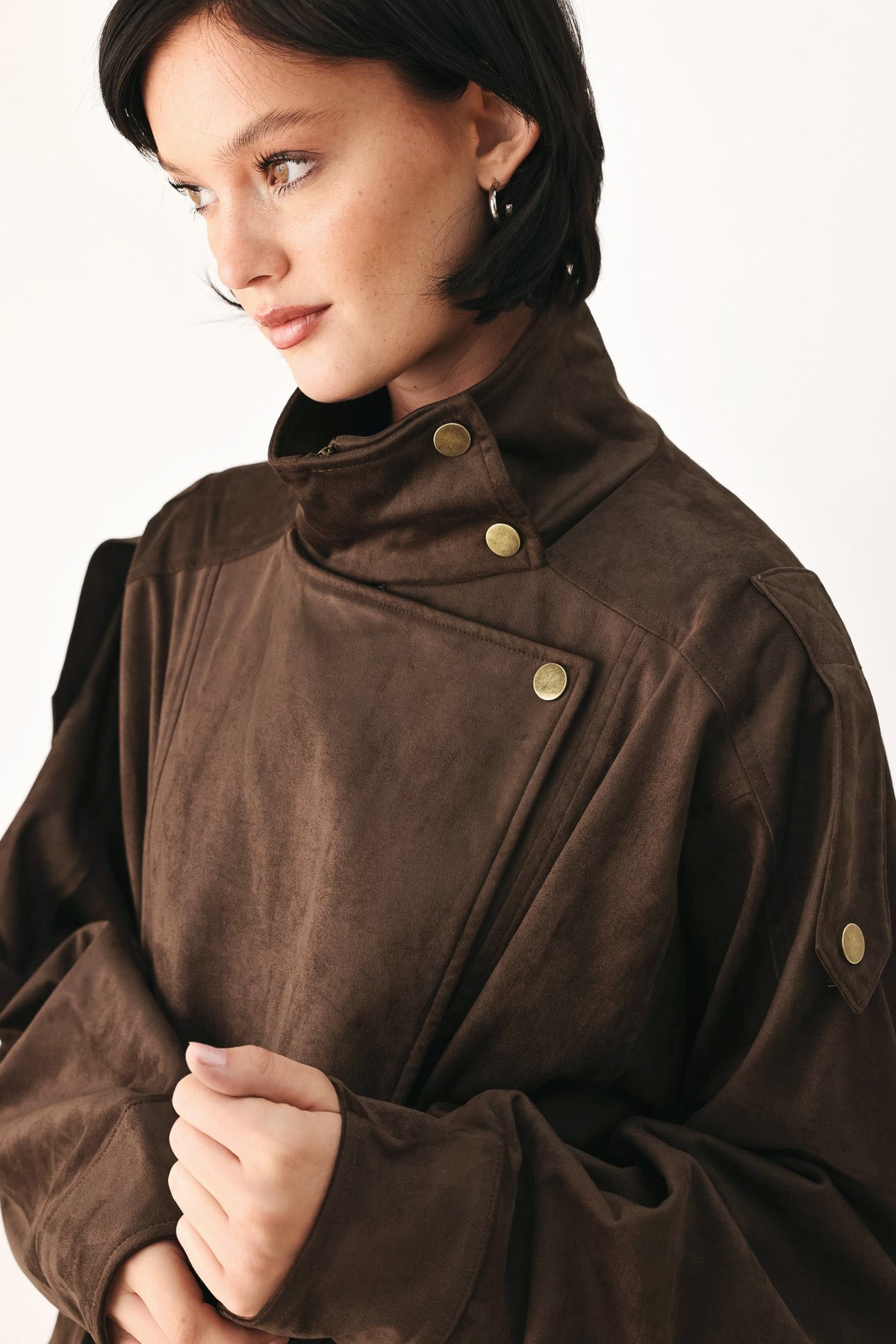A close-up of a model wearing a dark brown suede-like jacket with a high collar and brass snap buttons, set against a plain light background.
