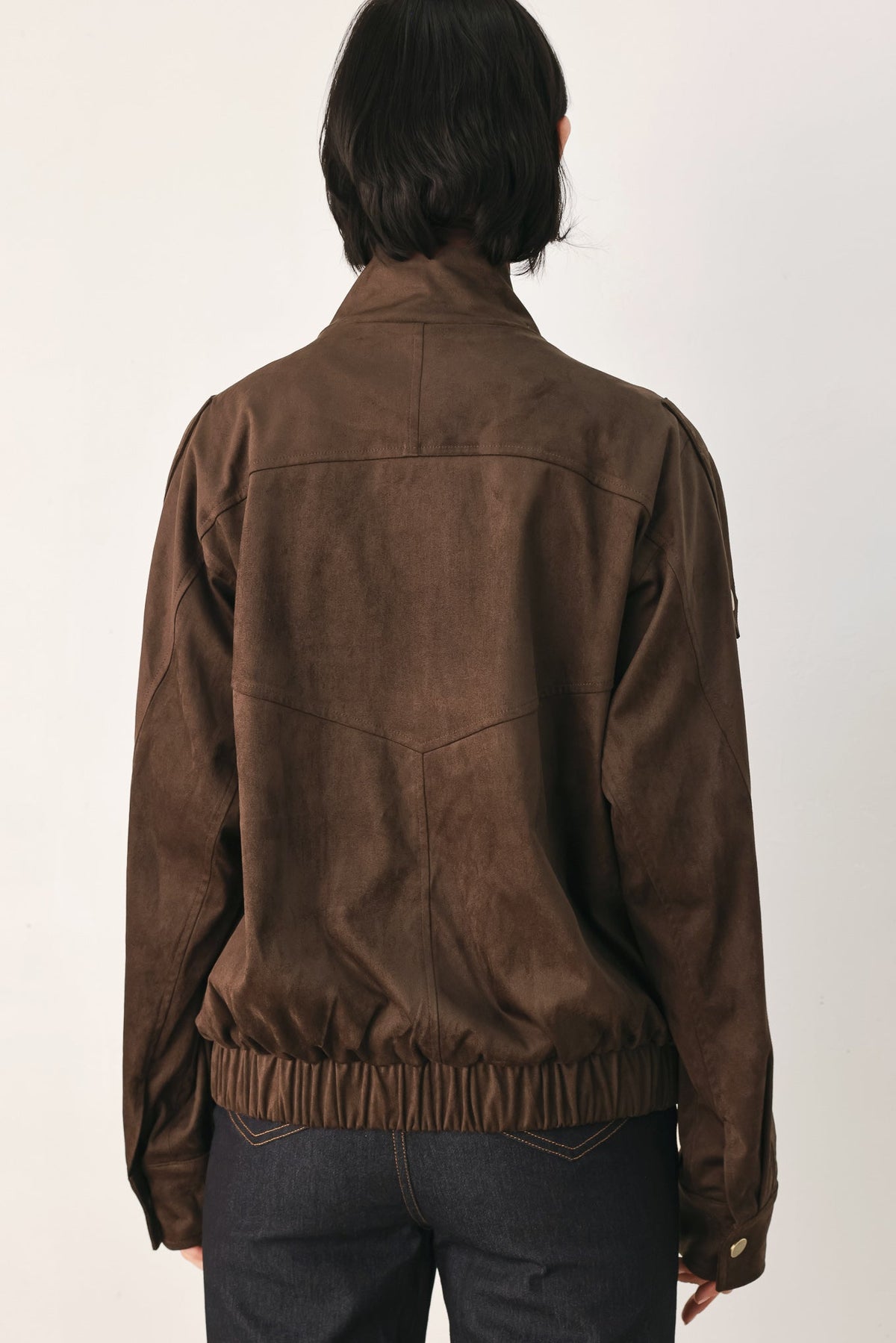 A person viewed from the back wearing a brown suede or faux-suede jacket with a gathered elastic hem, paired with dark jeans, against a plain light background.