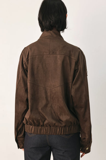 A person viewed from the back wearing a brown suede or faux-suede jacket with a gathered elastic hem, paired with dark jeans, against a plain light background.