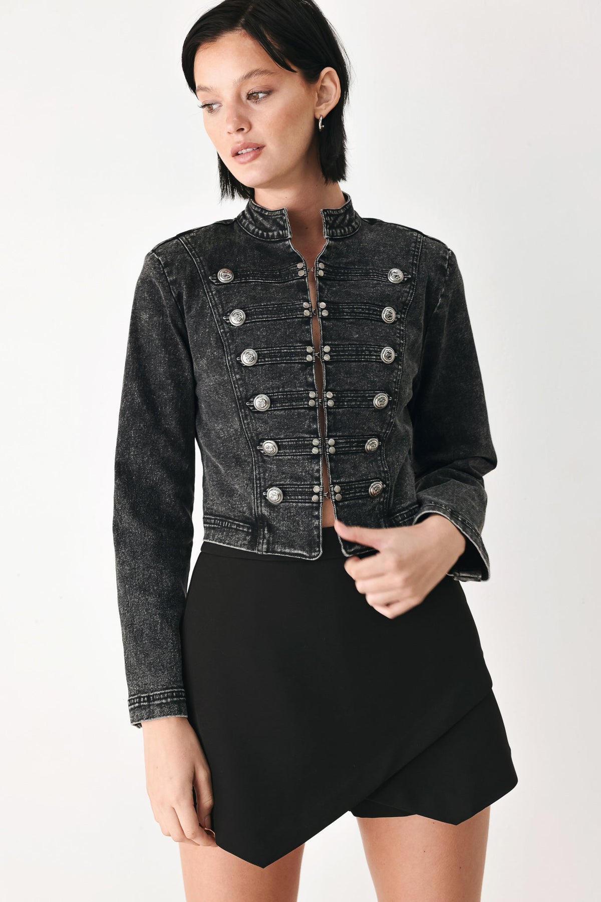 A model wearing a cropped black washed denim jacket with double-breasted silver buttons and a black asymmetrical mini skirt, photographed against a plain light gray studio background.