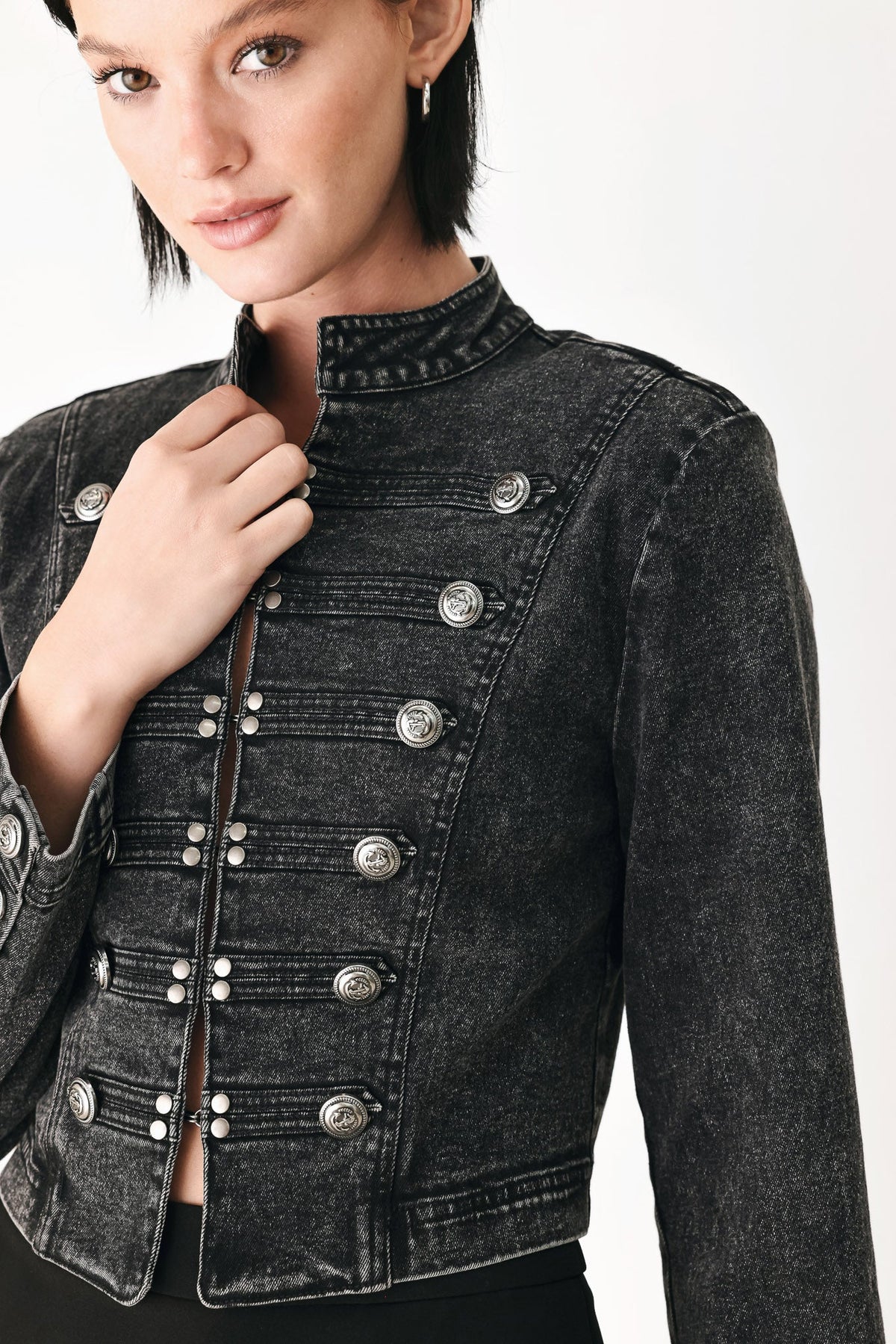 A close-up of a model wearing a cropped black denim jacket with silver military-style buttons and a high stand-up collar, set against a plain white background.