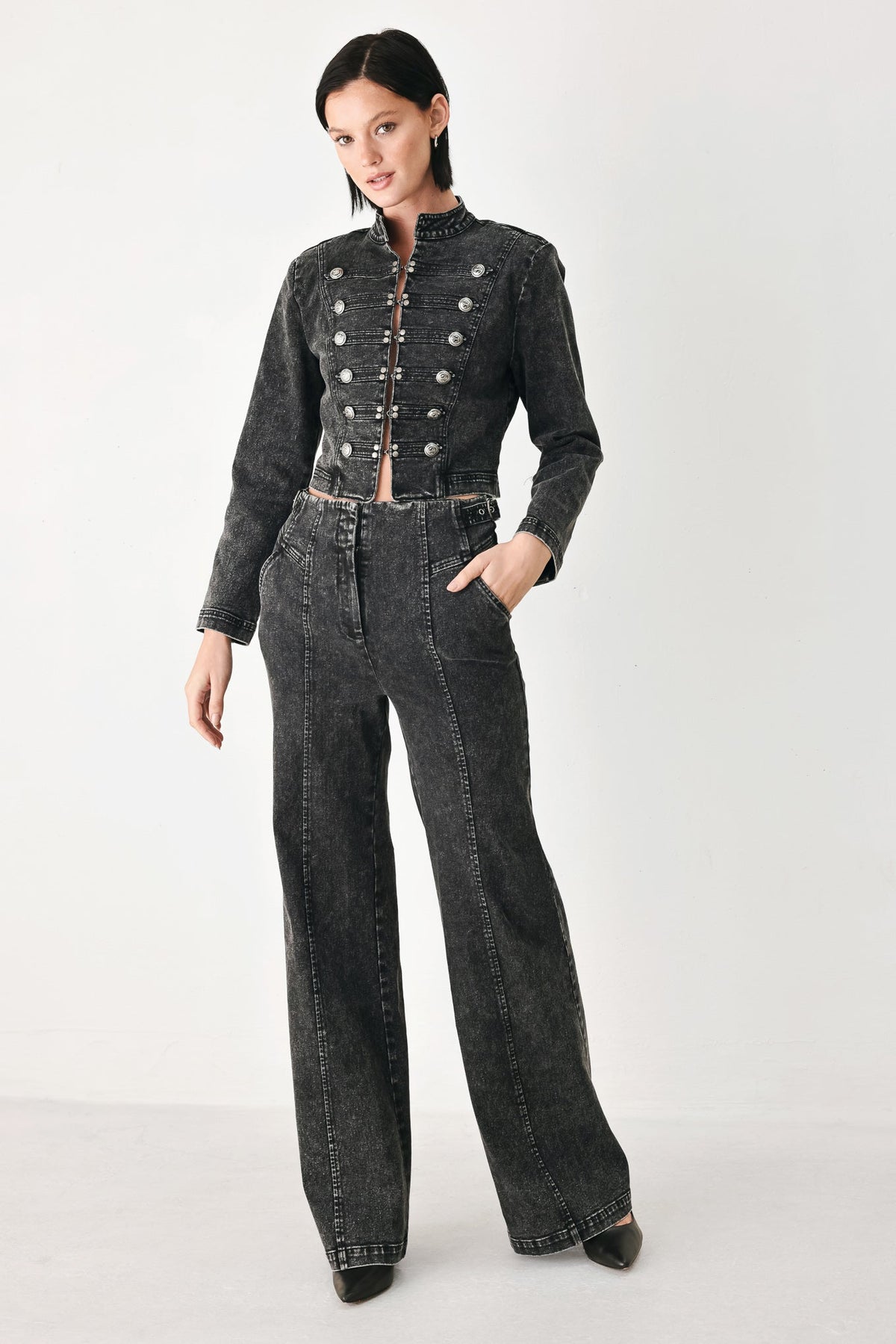 A model wearing a dark gray washed denim set consisting of a cropped, double-breasted jacket with silver buttons and matching wide-leg jeans, photographed against a plain light gray studio background.