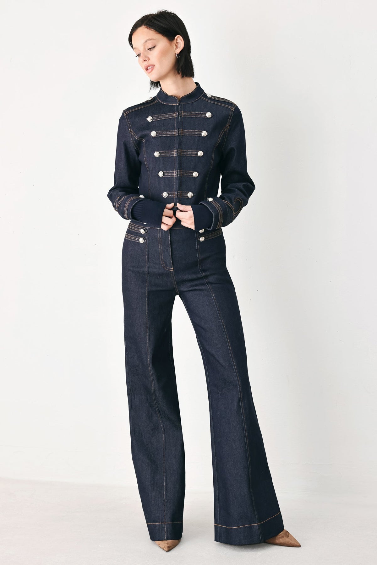 A model wearing a dark navy blue denim jumpsuit with a double-breasted military-style design, featuring silver buttons and contrast stitching, set against a plain light gray studio background.